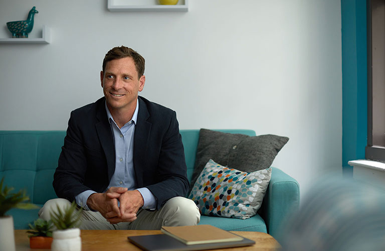 Lockwood Holmes Jr., wearing a blue sports coat and a broad smile, sitting on a teal couch with his hands clasped together.